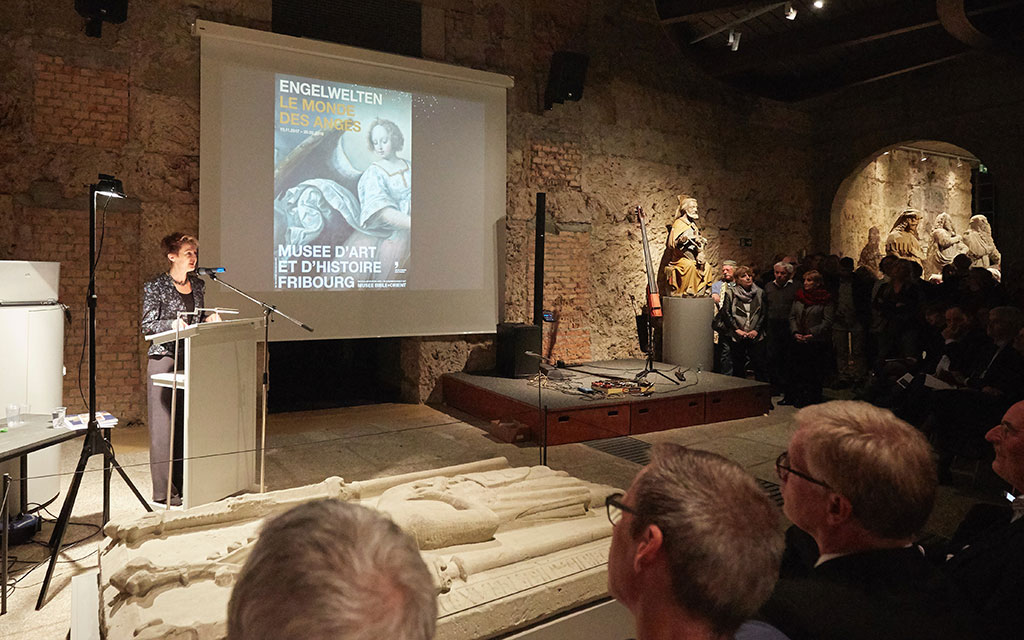 La consigliera federale Simonetta Sommaruga sul podio mentre parla a un microfono. Sullo sfondo si vede il manifesto della mostra “Engelwelten - Le monde des anges” del Museo d’arte di Friburgo.