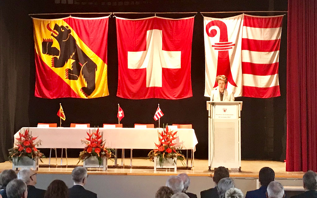 La consigliera federale Simonetta Sommaruga in tribuna al microfono, dietro di lei le bandiere del Canton Berna, della Svizzera e del Canton Giura.